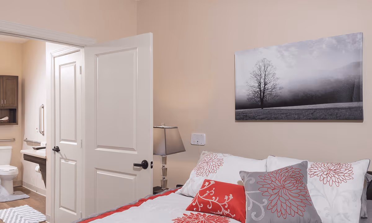 A bedroom with a bed featuring white and red floral pillows and bedding. A bedside table with a lamp is next to the bed. On the wall above the bed is a black and white picture of a tree in a foggy landscape. An open door reveals a bathroom with a toilet, sink, and grab bars.