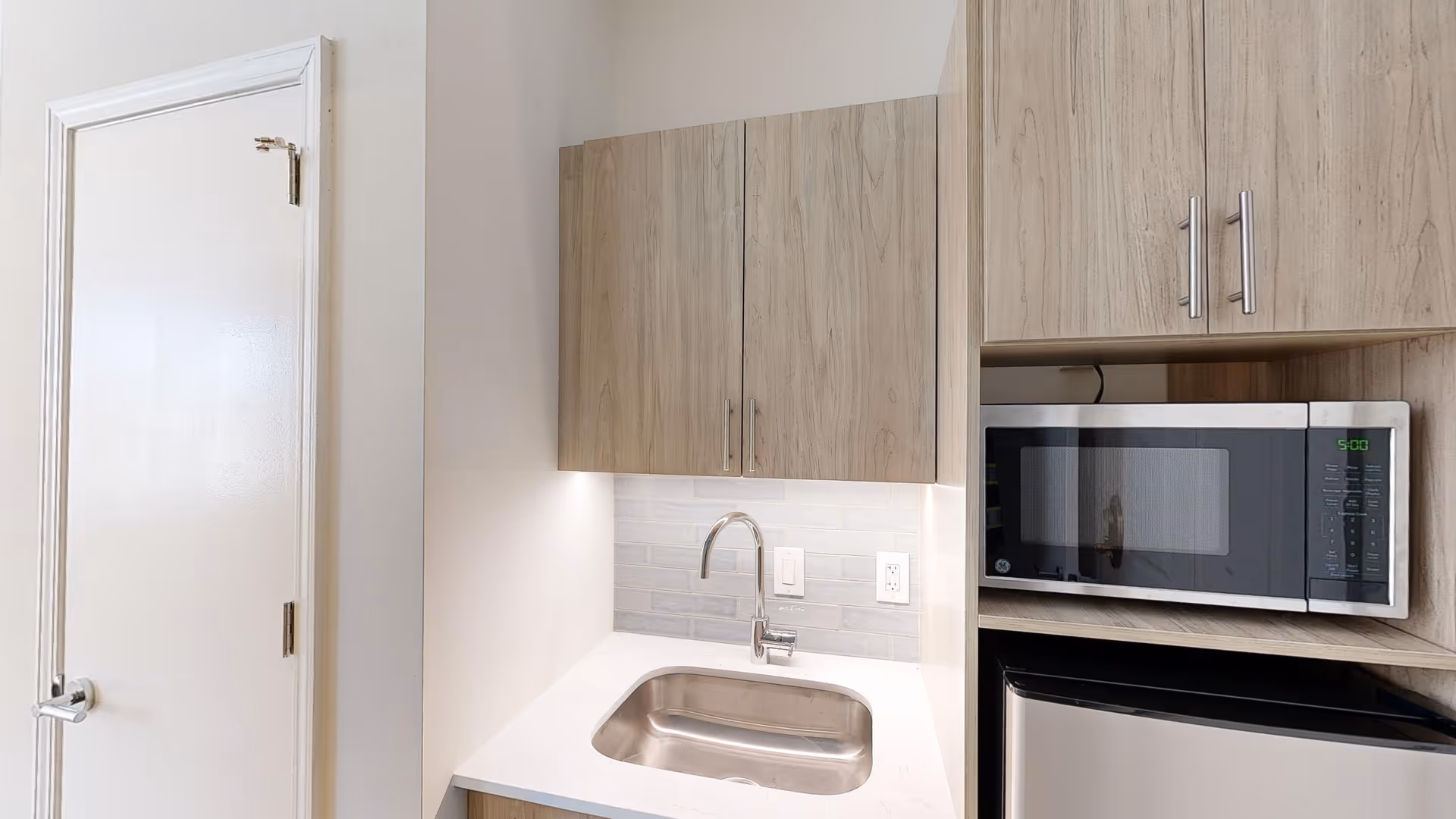 A small kitchenette area featuring a stainless steel sink with a modern faucet, light wood cabinets above and beside the sink, a microwave oven, and a mini refrigerator. The backsplash is made of light gray tiles, and there is a white door to the left of the kitchenette.
