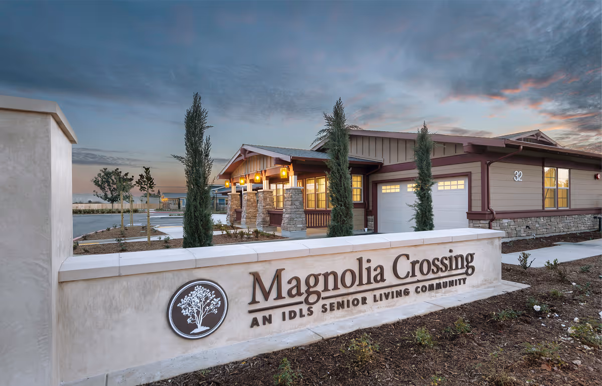 Exterior view of Magnolia Crossing Assisted Living facility at dusk, showing the building with warm lights on, a stone and stucco sign with the facility name, and landscaped surroundings.