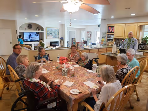 A group of elderly people sitting around a large dining table in a well-lit room. The table is covered with a patterned tablecloth and has a centerpiece with flowers. Some individuals are in wheelchairs, and others are seated in wooden chairs. In the background, there is a kitchen area with wooden cabinets and a man standing near the counter. A ceiling fan with lights is visible above the table, and a television and other furniture can be seen in the adjacent living space.