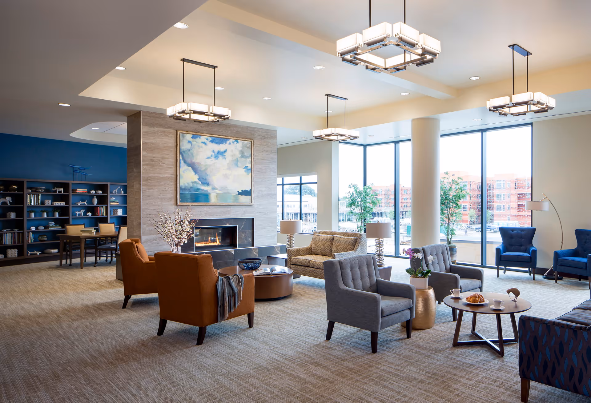 A spacious and well-lit living room area in a senior living facility with large windows letting in natural light. The room features a modern fireplace with a painting above it, several comfortable chairs and sofas arranged around coffee tables, and decorative plants. In the background, there is a bookshelf and a table with chairs, creating a cozy and inviting atmosphere.