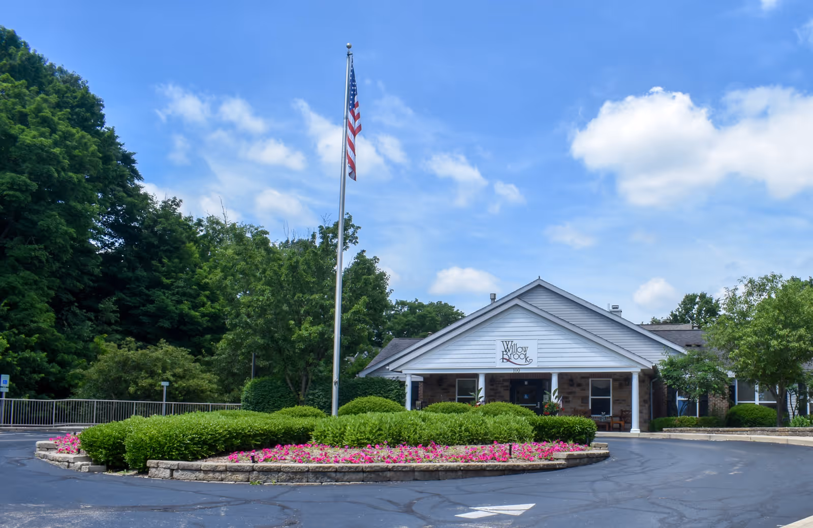 Front entrance of Willow Brook Christian Village showing the building facade, an American flag on a tall flagpole, and a landscaped circular driveway with flowers.