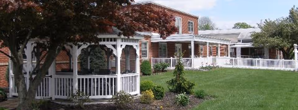 A red brick building with white trim and a white gazebo in the foreground surrounded by green grass, shrubs, and trees under a partly cloudy sky.