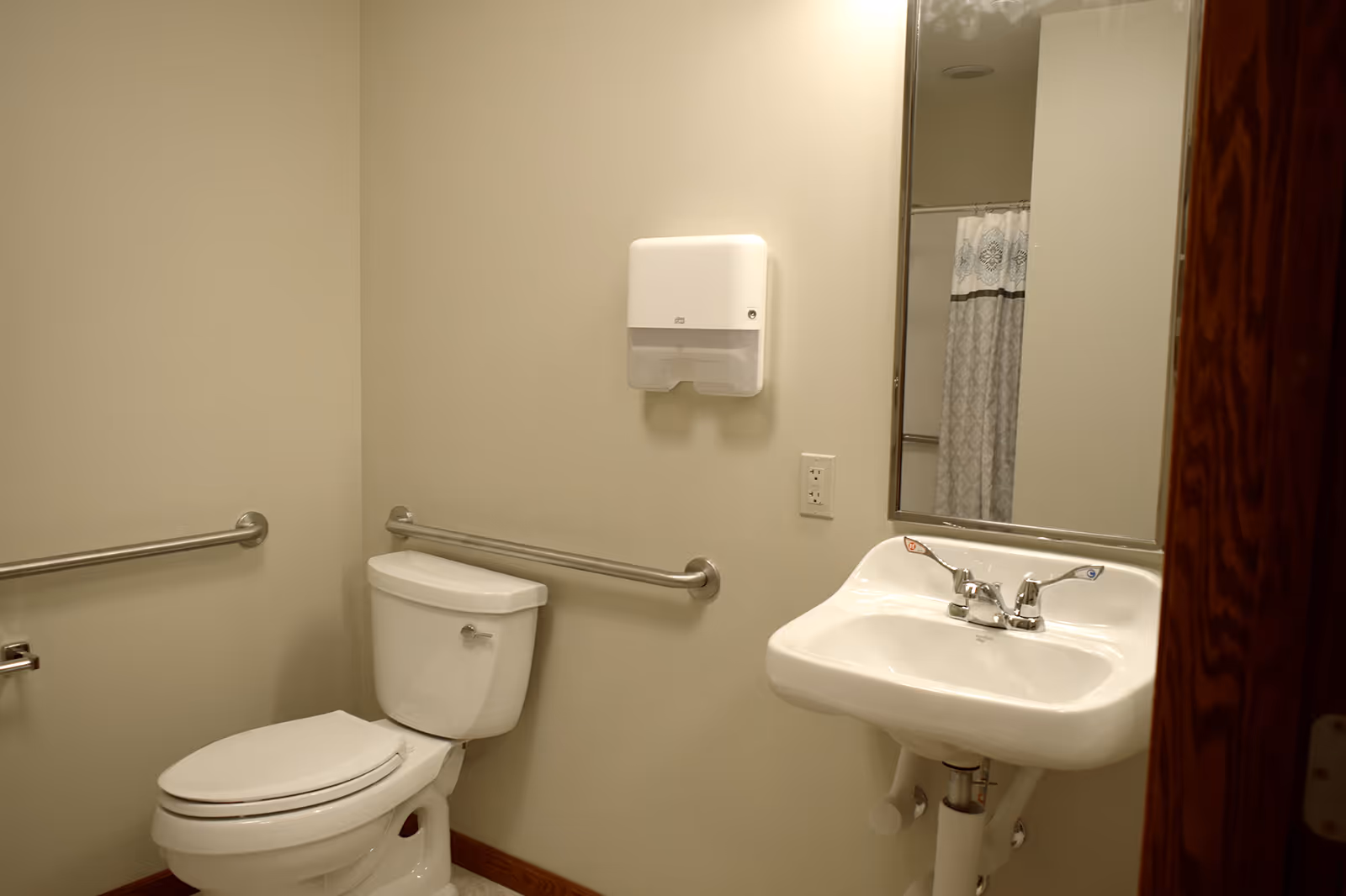 Accessible bathroom with a toilet, wall-mounted grab bars, a sink and mirror, and a paper towel dispenser.