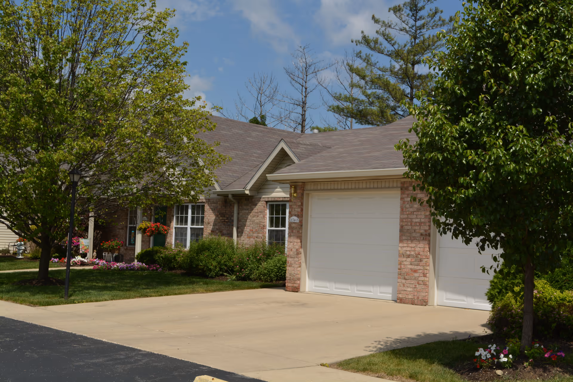 Exterior view of a single-story brick residential building with a two-car garage, surrounded by green trees and shrubs under a partly cloudy sky.