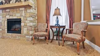 A cozy sitting area with two upholstered armchairs and a small wooden table with a decorative lamp between them, positioned in front of a stone fireplace. The room has beige carpet and warm-toned walls with a window covered by curtains behind the chairs.