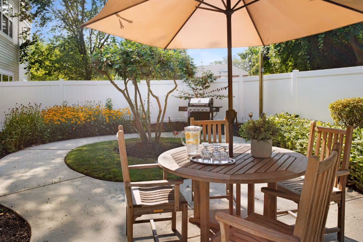 Outdoor patio area with a round wooden table and four wooden chairs under a large beige umbrella. On the table is a tray with a pitcher of lemon water and four glasses, along with a potted plant. The patio is surrounded by a white fence, green bushes, flowering plants, and a barbecue grill in the background.
