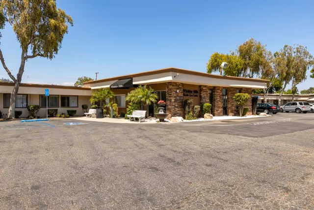 Exterior view of Whittier Hills Healthcare Center showing a single-story building with stone accents, a black awning over the entrance, several trees, bushes, benches, and a parking lot with a few cars and handicap parking spaces.