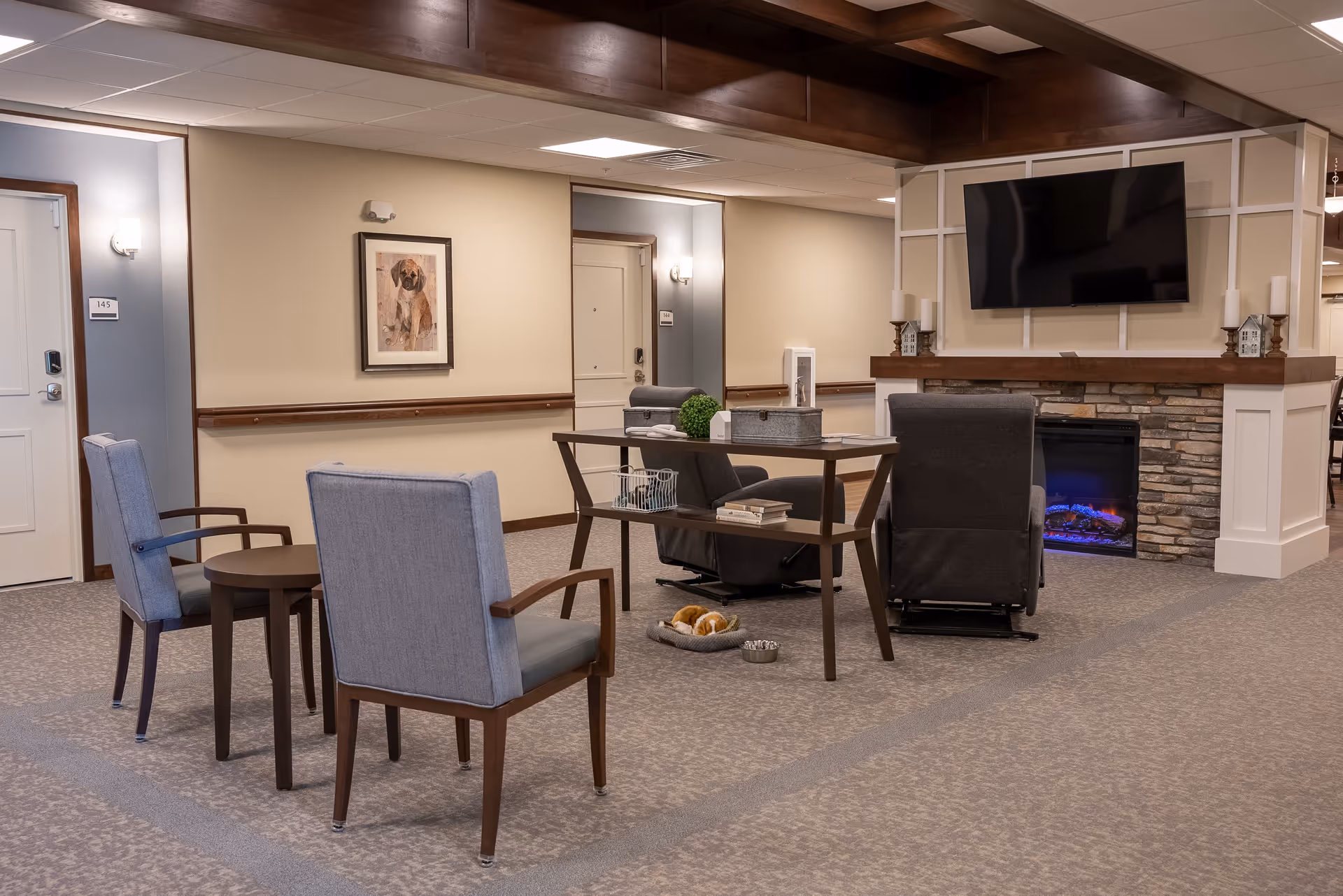 A cozy senior living common area with two gray armchairs and a small round table, a wooden table with books and decorative items, two dark recliners facing a stone fireplace with a mounted flat-screen TV above it. The walls are beige with wooden trim, and there is a framed picture of a dog on the wall. The carpet is patterned in neutral tones.