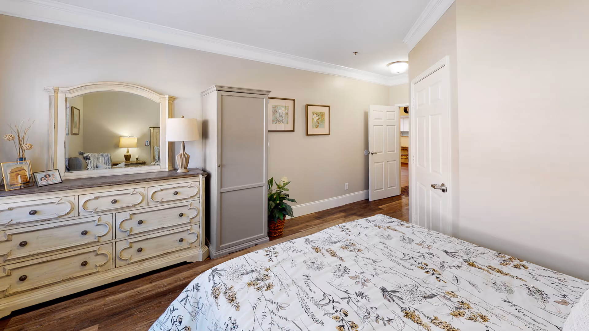 A bedroom with a floral patterned bedspread, a distressed white dresser with a large mirror, a table lamp, framed photos, a gray wardrobe, two framed floral pictures on the wall, and an open door leading to another room.