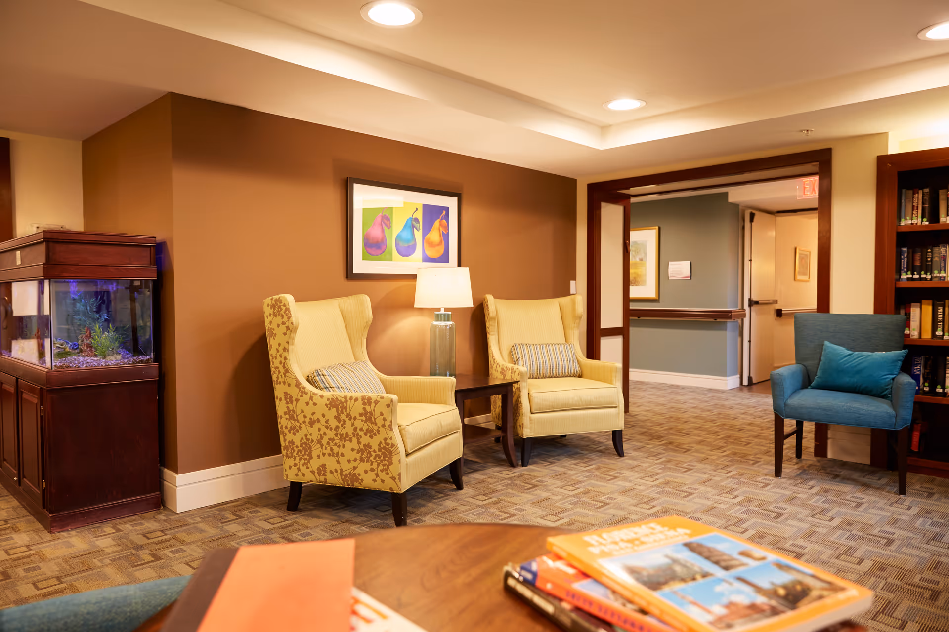 A cozy seating area in an assisted living facility featuring two yellow patterned armchairs with striped cushions, a small wooden side table with a lamp, a blue armchair with a blue cushion, a large fish tank on a wooden cabinet, and a bookshelf filled with books. The walls are painted in warm tones with framed artwork, and the floor is carpeted with a geometric pattern.