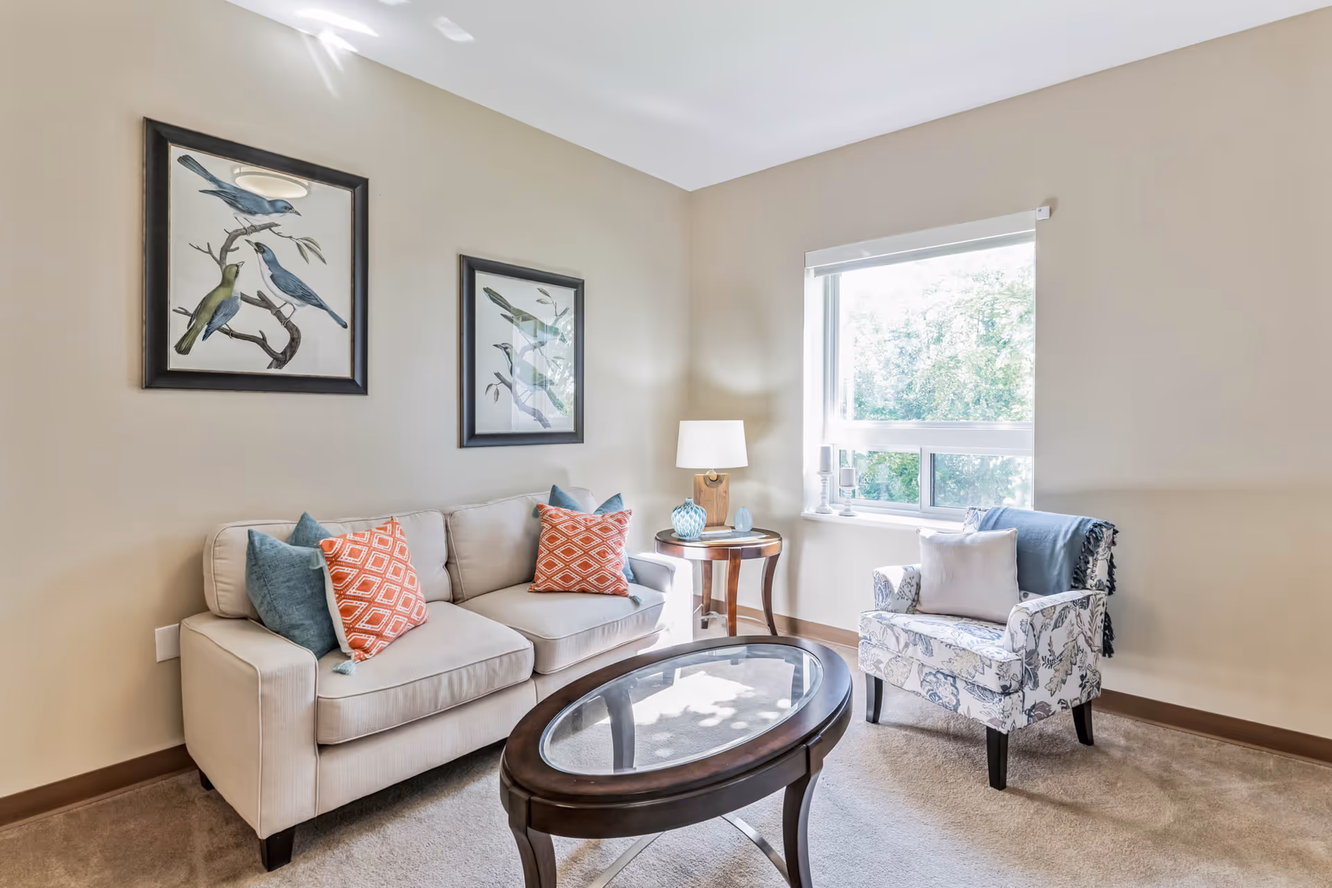 Bright living room with a beige sofa, patterned armchair, oval glass-top coffee table, bird artwork on the wall, and a window letting in natural light.