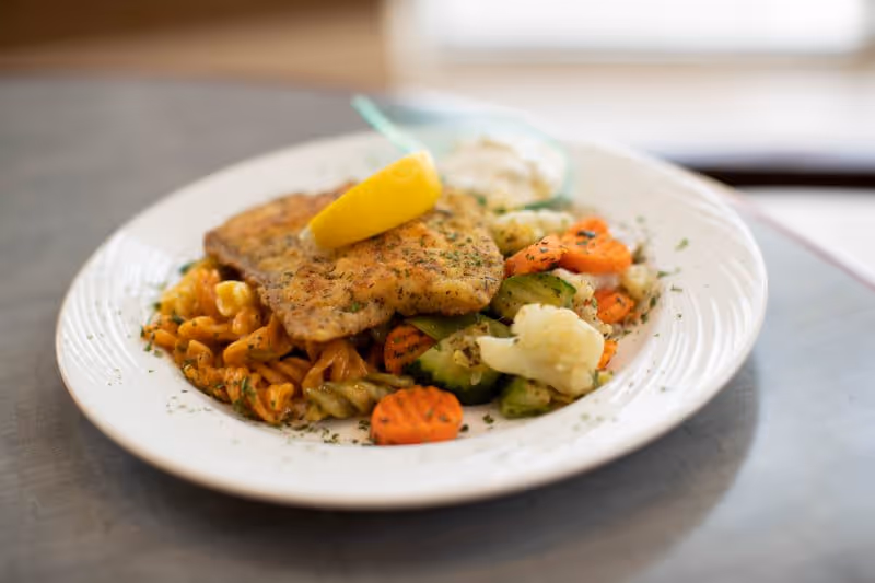 A plate of food featuring a seasoned and cooked fish fillet topped with a lemon wedge, served with tri-color rotini pasta and mixed steamed vegetables including carrots, cauliflower, and zucchini, with a small portion of sauce on the side.