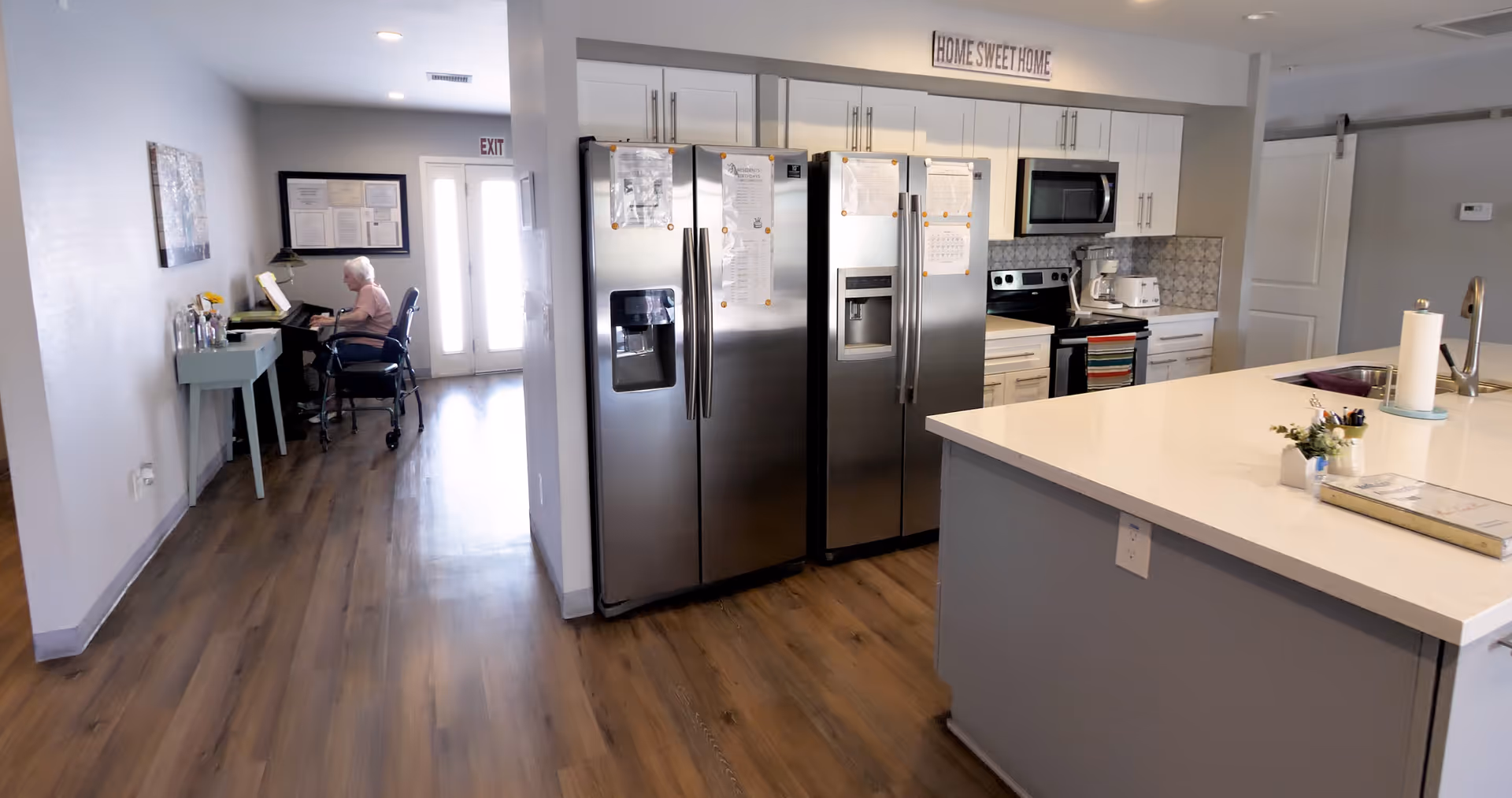Interior view of a senior living facility kitchen area with two stainless steel refrigerators, white cabinets, a stove, microwave, and a large kitchen island with a sink and paper towel holder. In the background, an elderly person is seated at a piano in a hallway with wooden flooring and a 'Home Sweet Home' sign above the kitchen cabinets.