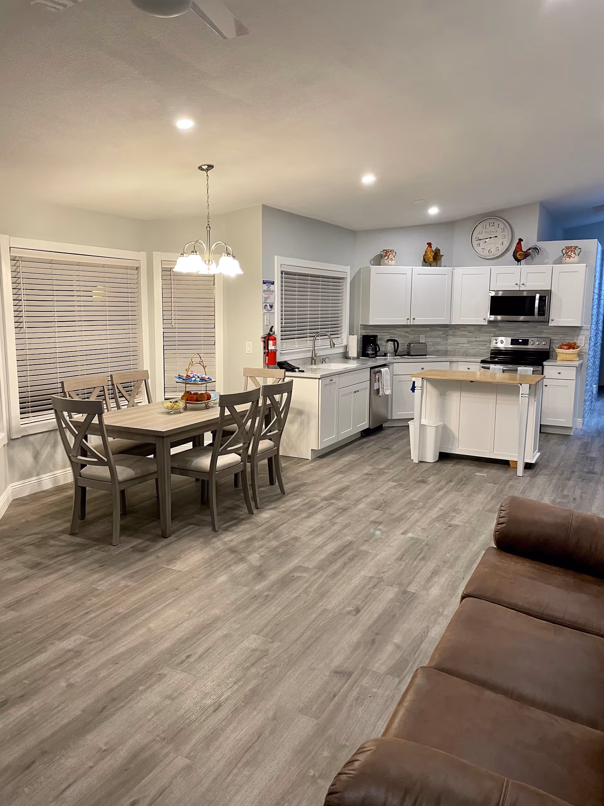 A modern open-concept kitchen and dining area with light gray wood flooring. The kitchen features white cabinets, stainless steel appliances including a microwave and stove, a small island with a wooden countertop, and a window with blinds above the sink. The dining area has a wooden table with six chairs and a hanging light fixture above it. A brown leather sofa is partially visible in the foreground.