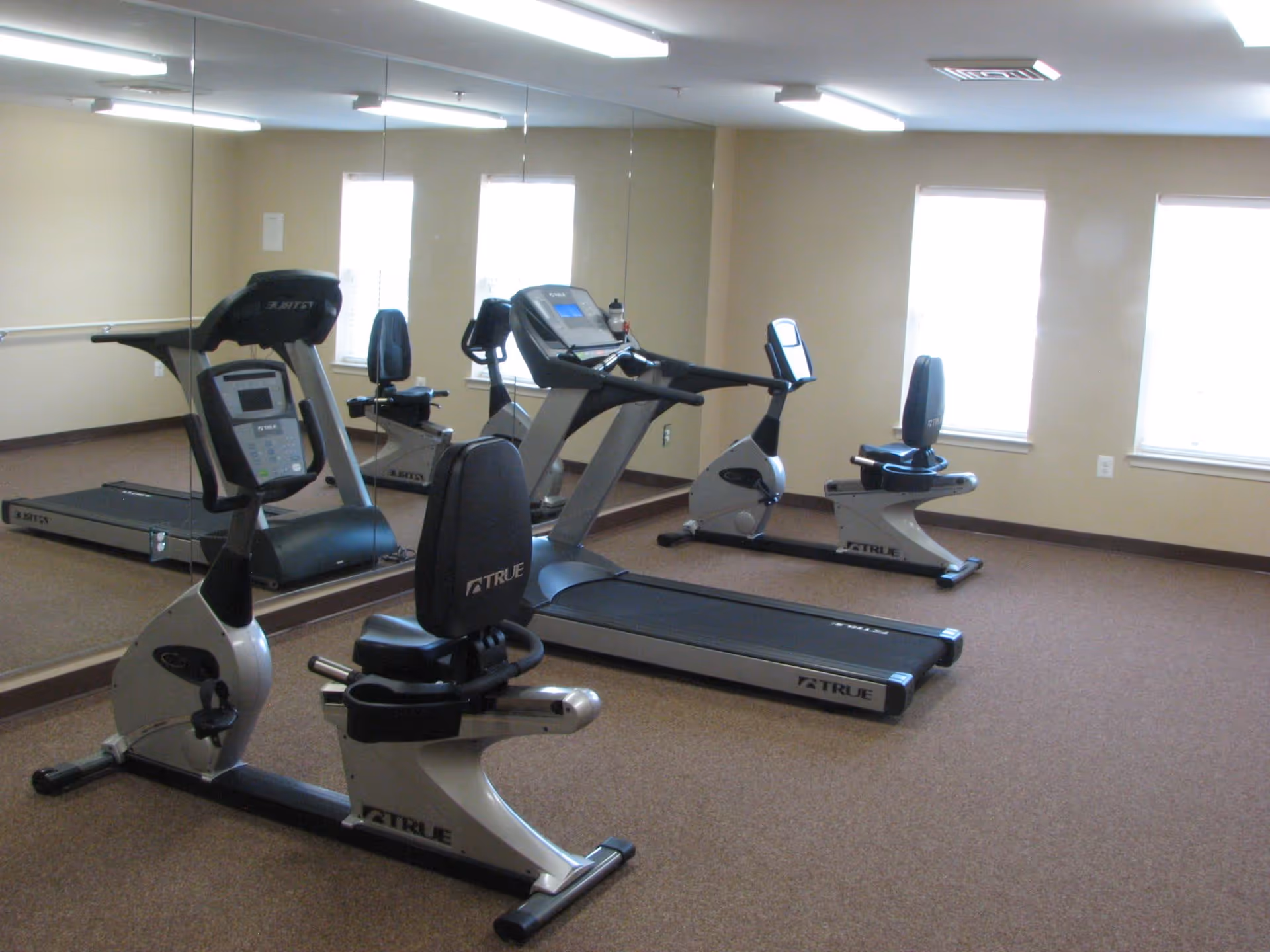 A small fitness room with exercise equipment including a treadmill and two stationary bikes. The room has beige walls, a brown carpet, three windows letting in natural light, and a large wall mirror reflecting the equipment.
