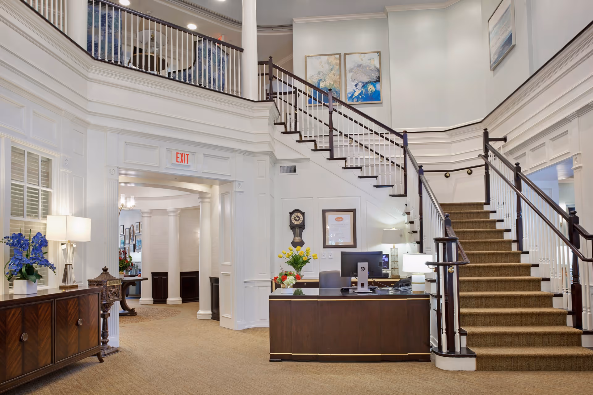 Interior view of a senior living facility lobby with a wooden reception desk, a staircase with white railings and dark wood handrails, beige carpet, white paneled walls, and decorative paintings. There are flowers on the desk and a lamp on a side cabinet. An exit sign is visible above an archway leading to another room with white columns.