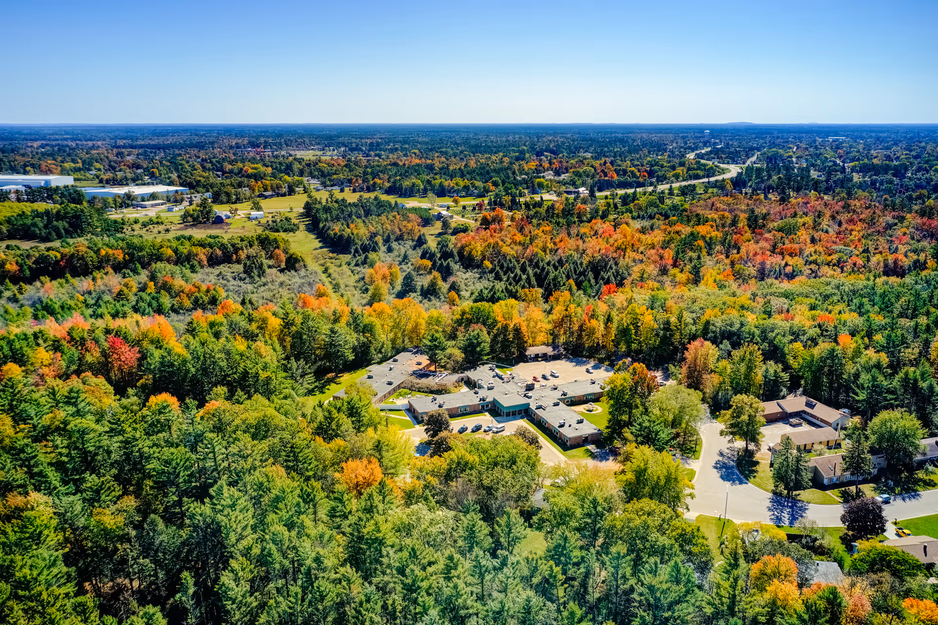 Aerial view of Edenbrook Wisconsin Rapids surrounded by dense forest with trees showing autumn colors. The facility buildings are visible in the center, with roads and parking areas nearby. The landscape extends to the horizon under a clear blue sky.