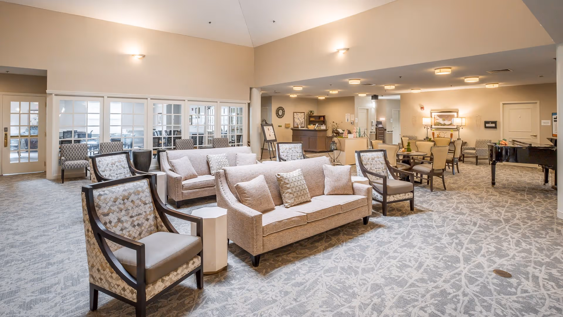 A spacious and well-lit senior living facility common area featuring multiple seating arrangements including beige sofas, patterned armchairs, and dining tables with chairs. The room has a light-colored carpet with a subtle pattern, a grand piano on the right side, and a reception desk towards the back. The walls are painted in neutral tones, and there are several light fixtures on the ceiling providing ample lighting.