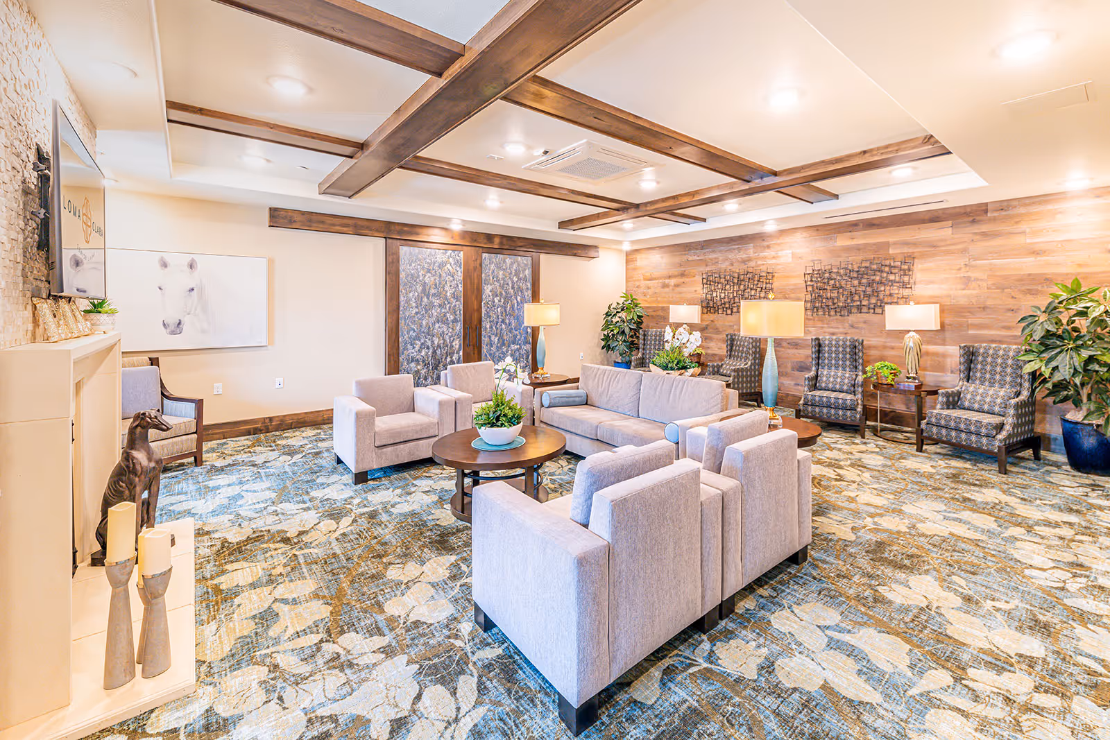 A bright and spacious senior living facility lounge with modern furniture including light gray armchairs and a sofa arranged around wooden coffee tables. The room features a patterned carpet, wooden accent walls, ceiling beams, several table lamps, potted plants, and decorative wall art. A fireplace with candles and a dog statue is visible on the left side, and a TV mounted above it displays the Loma Clara logo.
