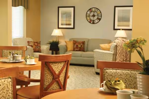 Cozy senior living lounge with sofas, chairs, tables, lamps, and framed wall art.