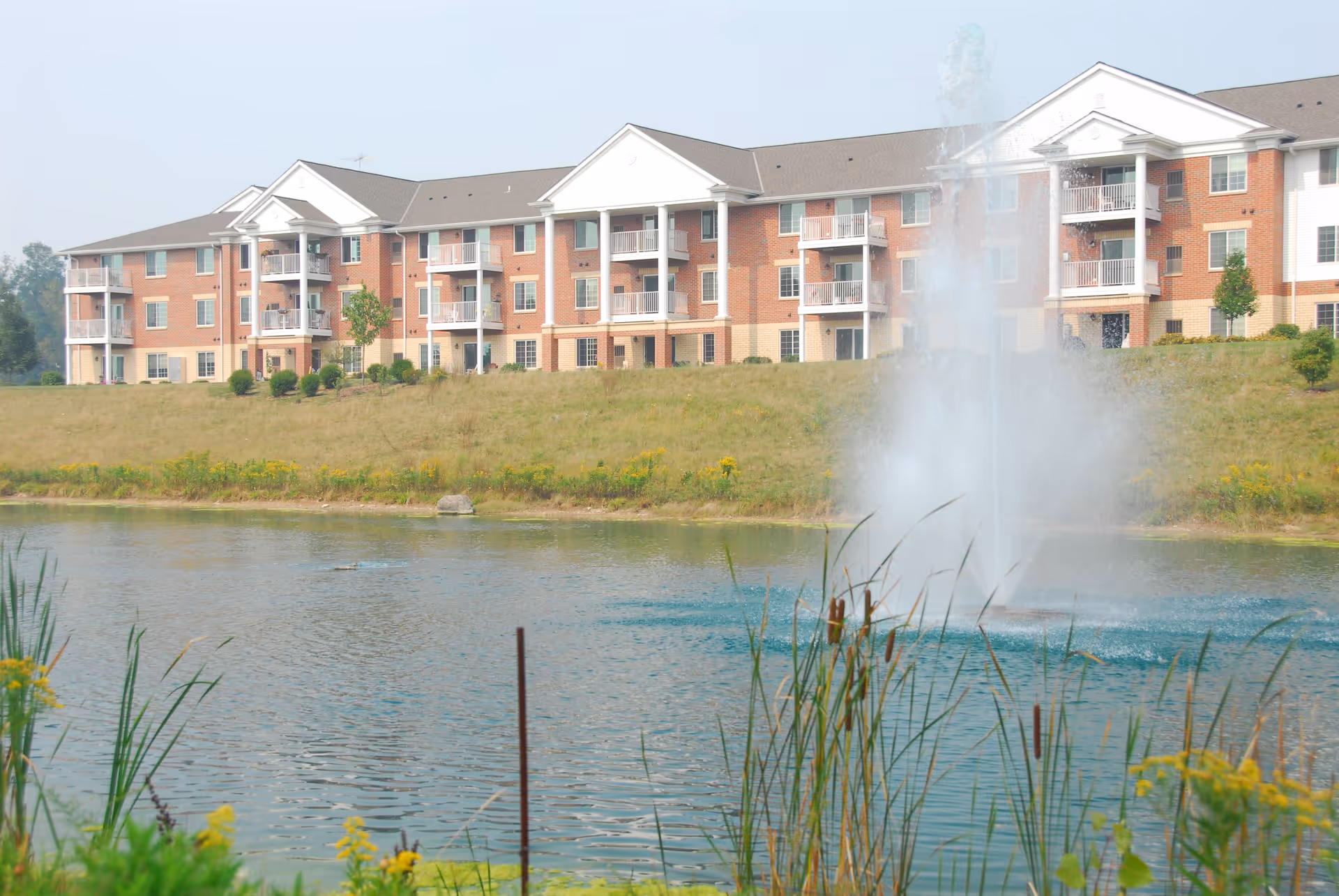 Three-story brick apartment building with balconies overlooking a pond and a fountain.