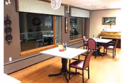 Small communal dining/activity room with tables and chairs, a piano, large windows, and wood flooring.