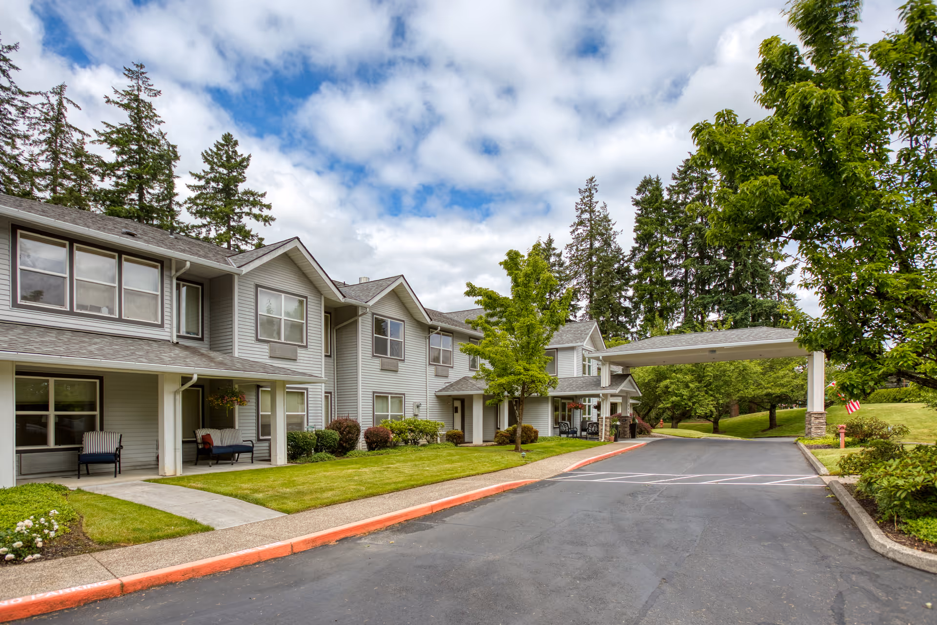Front exterior of a two-story assisted living building with a covered driveway entrance, lawn, and trees under a partly cloudy sky.