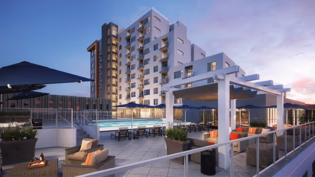 Outdoor rooftop patio area at sunset with lounge chairs, umbrellas, a swimming pool, and a pergola with cushioned seating. A multi-story building is visible in the background.