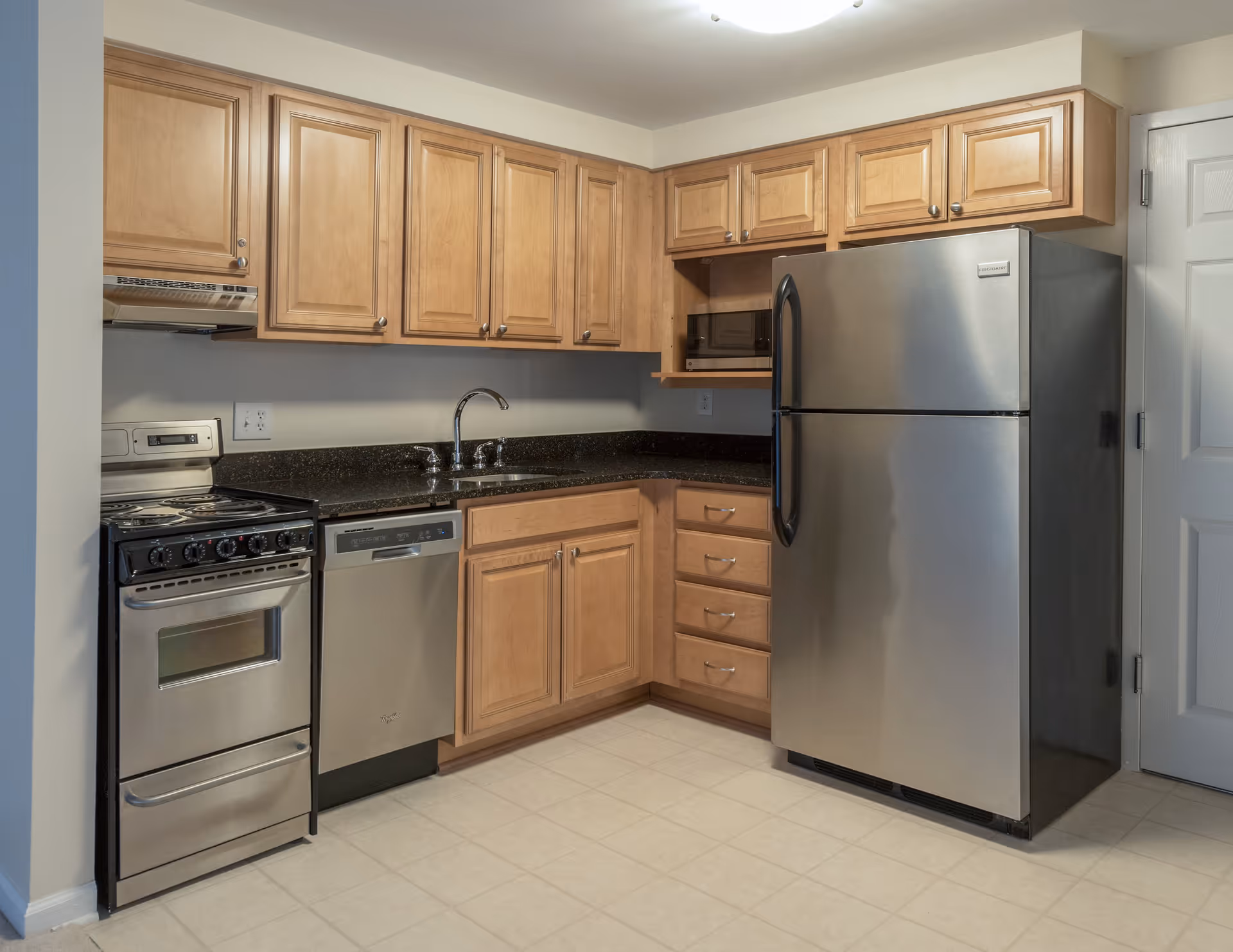 A compact kitchen with light wood cabinets, a stainless steel refrigerator, dishwasher, microwave, and an electric stove with oven. The countertops are dark granite, and the floor is covered with light-colored tiles.