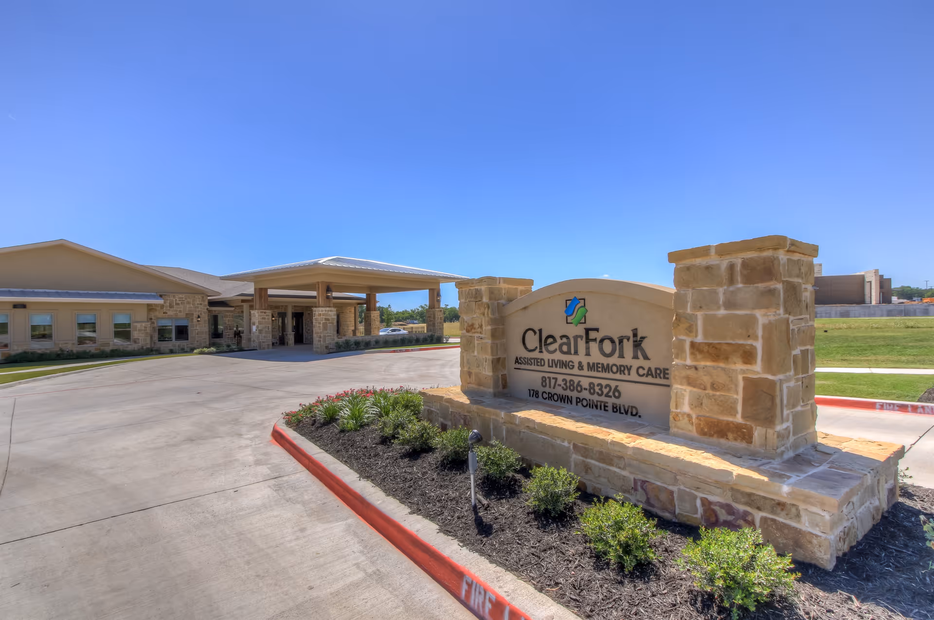 Exterior view of Clear Fork Assisted Living and Memory Care facility showing the entrance with a covered drop-off area and a stone sign with the facility name, phone number, and address. The sky is clear and blue, and there are some small bushes and plants around the sign.