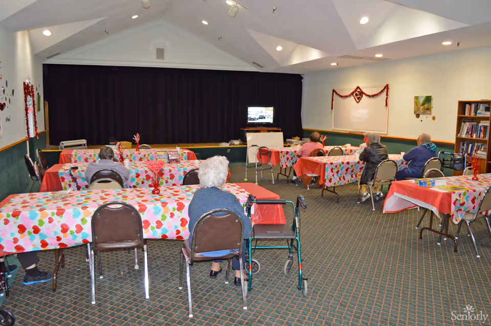 Residents seated at tables with heart-patterned coverings in a communal area of a senior living facility.