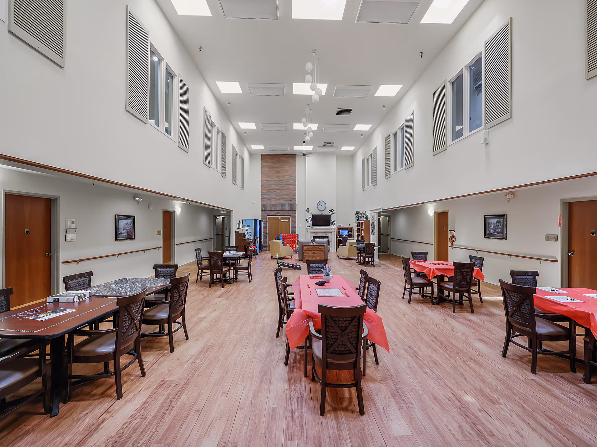 A spacious, well-lit common area in an assisted living facility with multiple tables and chairs arranged for dining or social activities. The room has high ceilings with skylights, light wood flooring, and white walls. There is a fireplace at the far end with a clock and TV mounted above it, and several doors along the side walls.