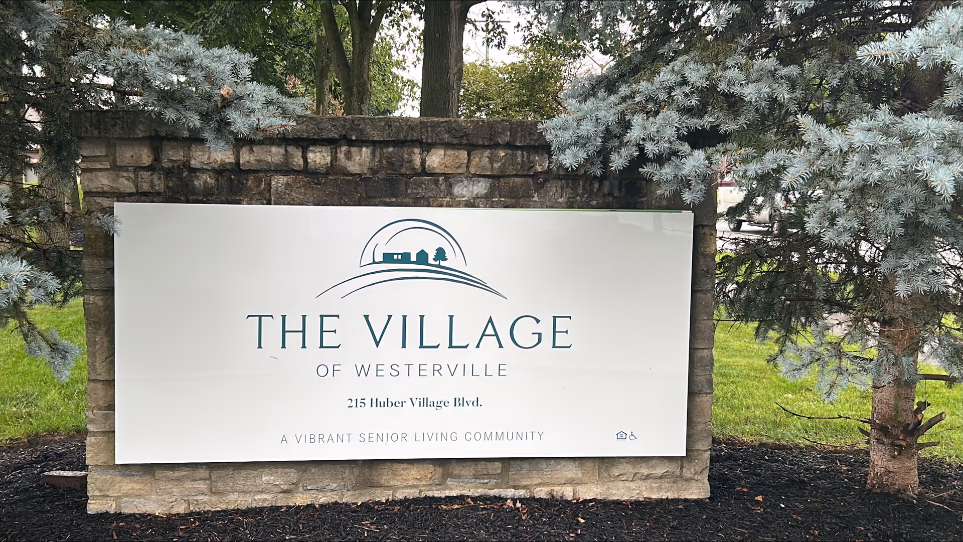 A stone sign at The Village of Westerville senior living community, surrounded by trees and grass. The sign displays the community name, address 215 Huber Village Blvd., and the tagline 'A Vibrant Senior Living Community.'