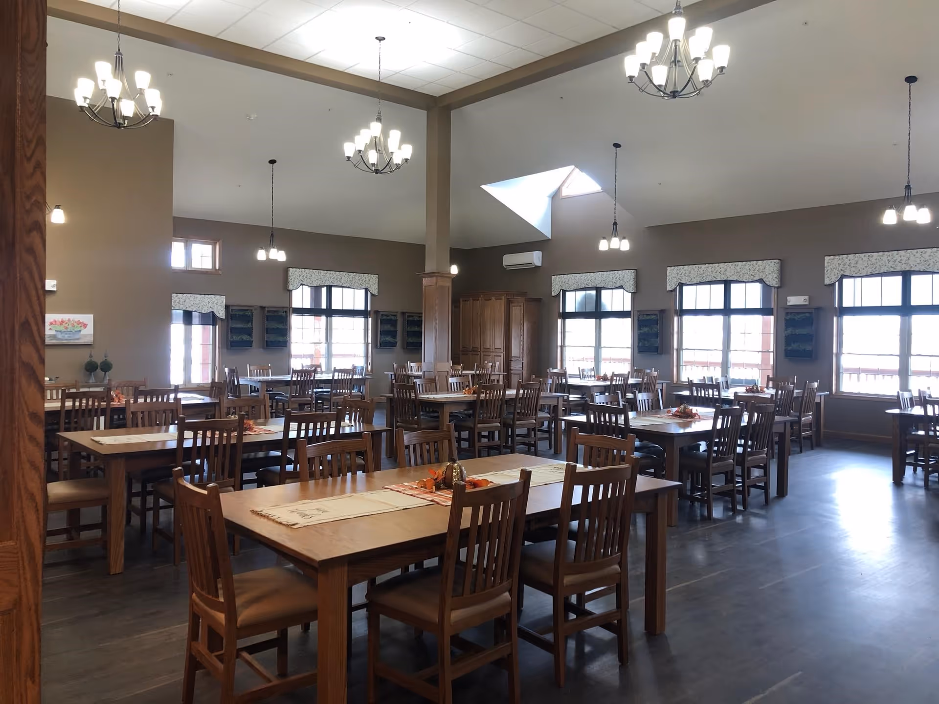 A spacious dining room with multiple wooden tables and chairs arranged neatly. The room features large windows with valances, high ceilings with chandeliers and pendant lights, and a wooden floor. The tables have placemats and small decorative centerpieces.