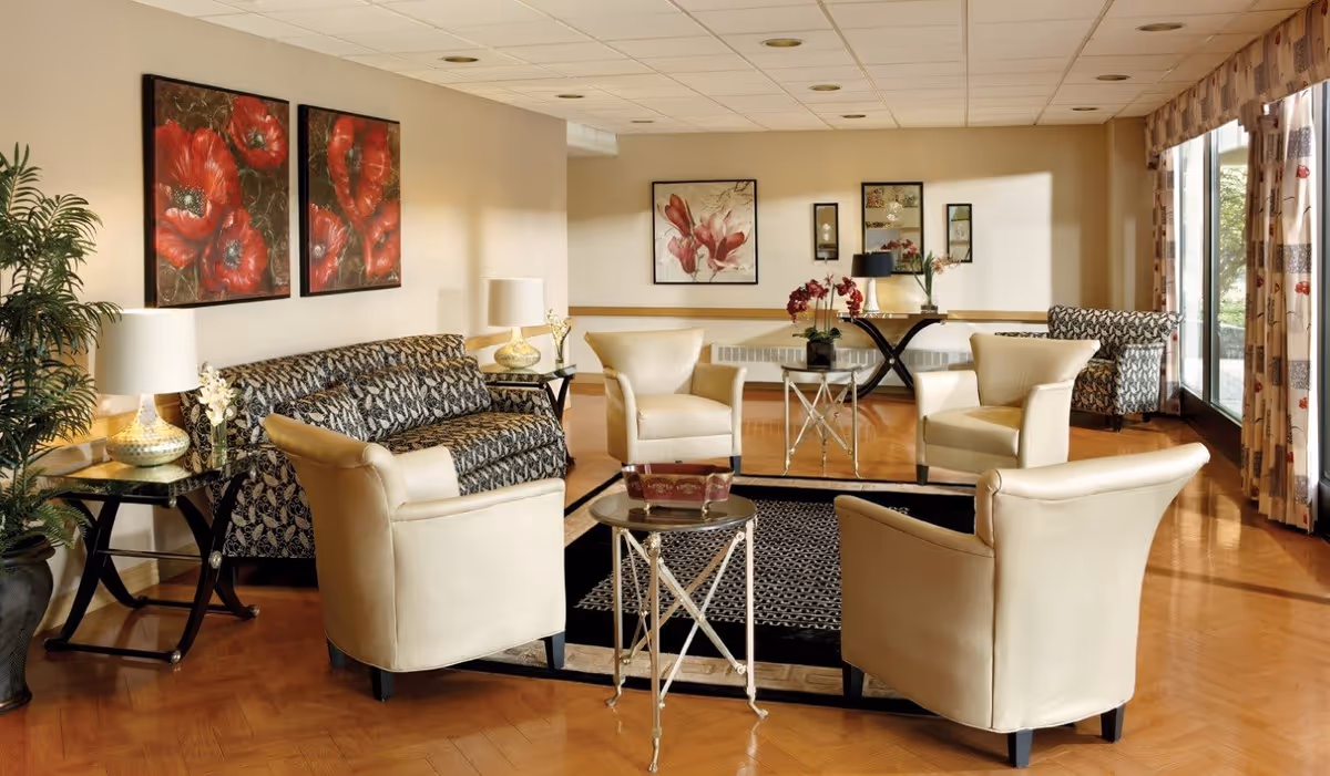 A well-lit living room area with four cream-colored armchairs arranged around a small round table on a patterned rug. Behind the chairs is a patterned sofa with two side tables holding lamps and flowers. The walls are decorated with floral paintings and mirrors. Large windows with patterned curtains allow natural light to fill the room.