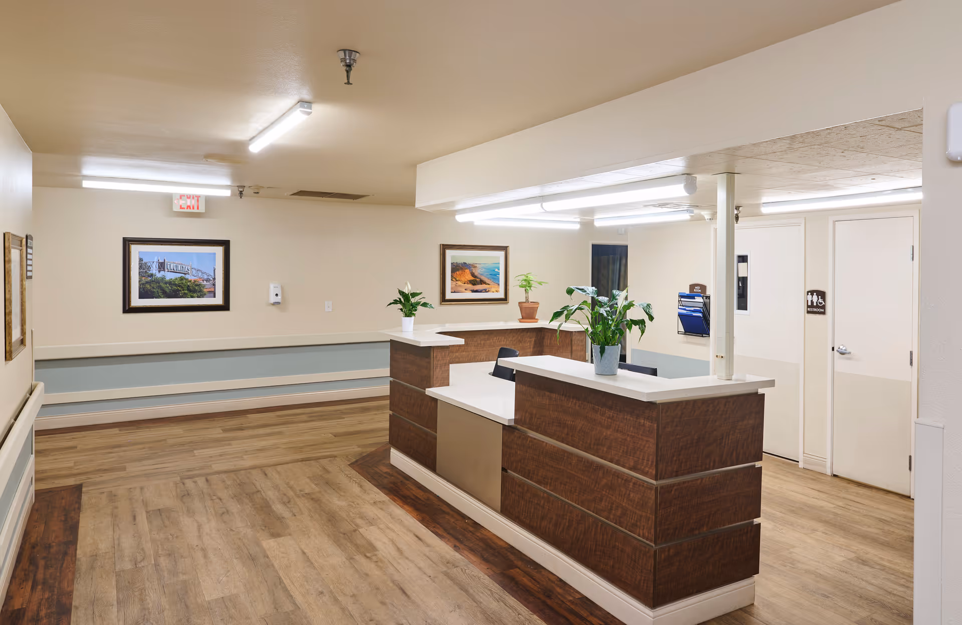 Reception area inside a nursing and rehabilitation center with a wooden front desk adorned with potted plants, light-colored walls, framed landscape pictures, wooden flooring, and fluorescent ceiling lights. There are doors to the right, one marked as a restroom.