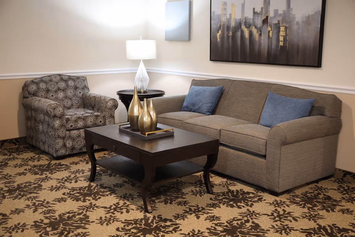 A cozy living room area with a patterned armchair, a gray sofa with two blue cushions, a dark wooden coffee table with three decorative gold vases, a side table with a white lamp, and a large abstract cityscape painting on the wall.