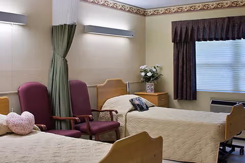 A nursing home room with two single beds covered in beige quilts, two maroon armchairs between the beds, a nightstand with a vase of white flowers and a framed photo, a window with dark curtains, and a wall-mounted light above the beds.