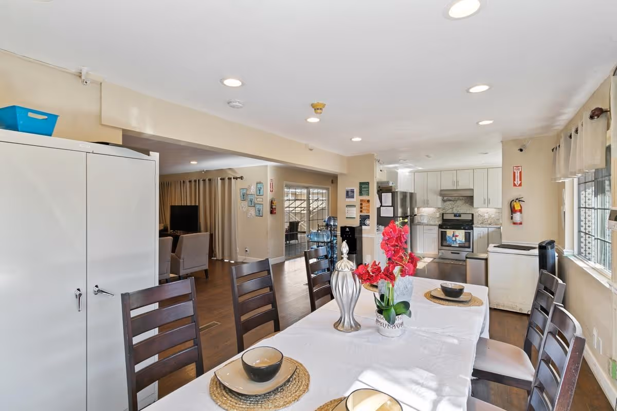 A dining table set with place settings and a vase of red flowers in a bright open dining area that connects to a kitchen and living room.