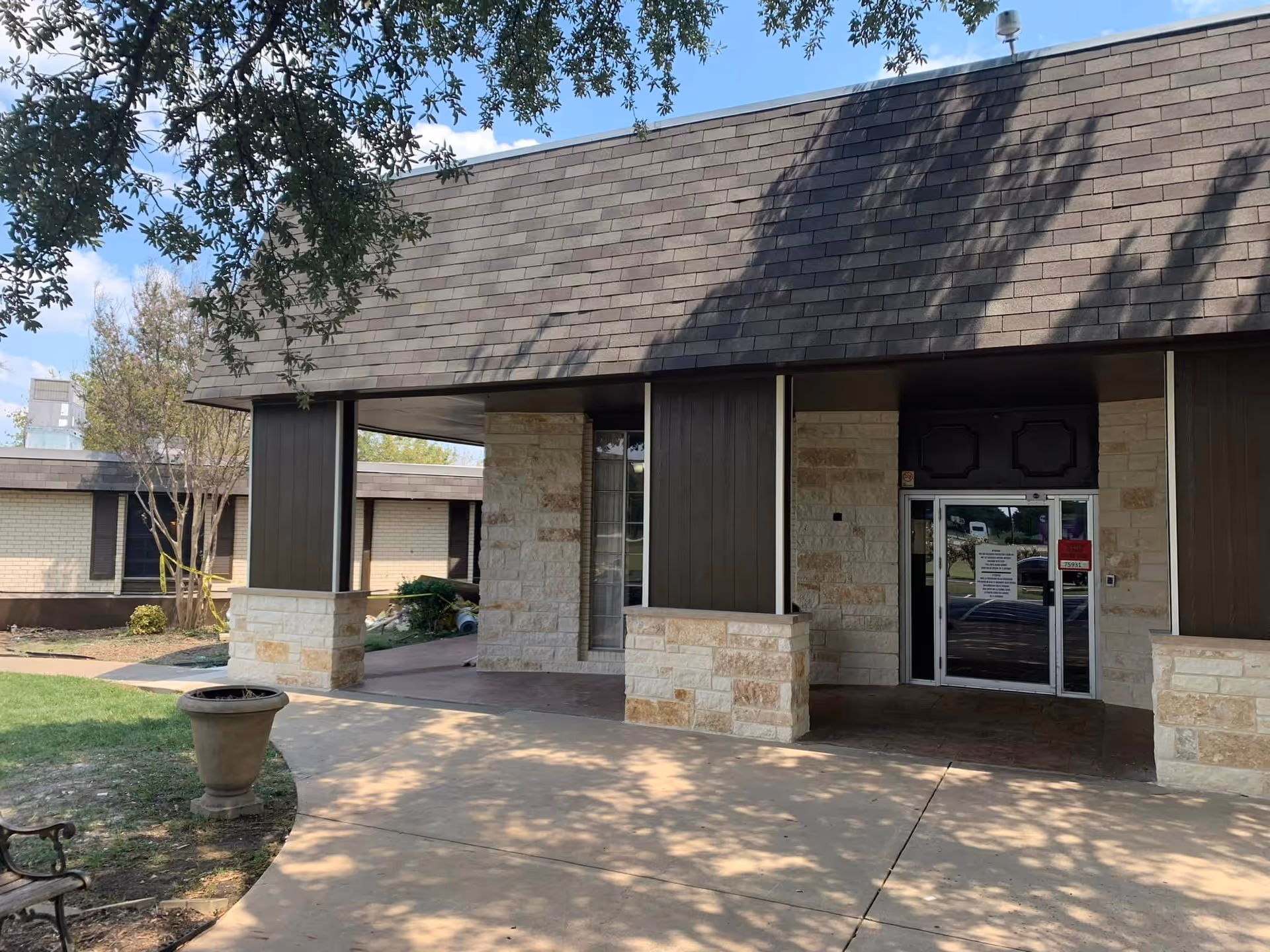 Entrance of North Park Health and Rehabilitation Center showing a covered walkway with stone pillars and a glass door entrance. There is a large tree casting shadows on the walkway and a planter on the left side.