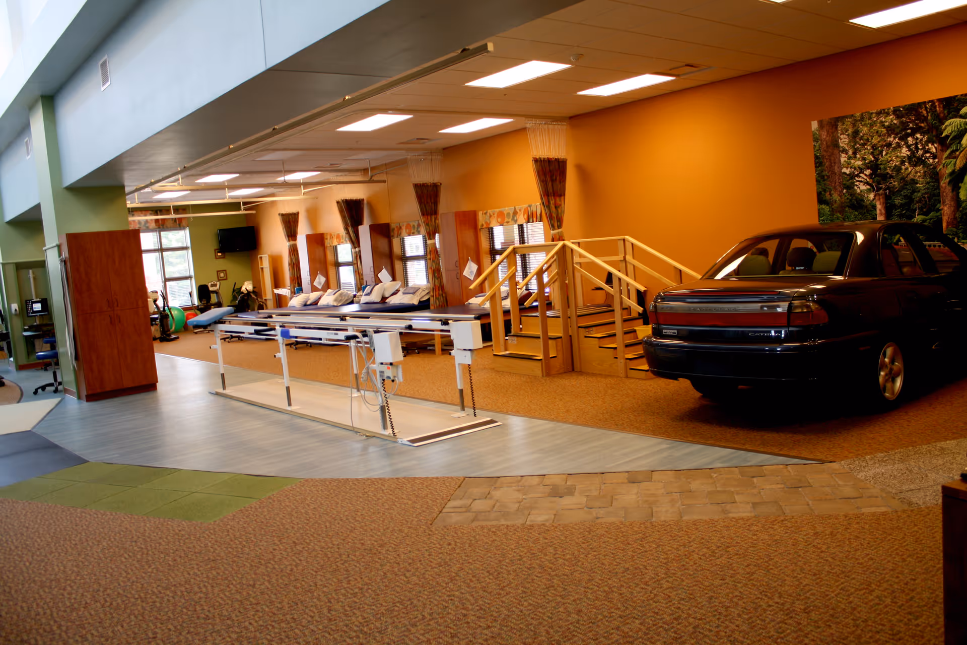 Interior view of a rehabilitation or physical therapy room in an assisted living facility, featuring parallel bars for walking exercises, a set of stairs with handrails for mobility training, several therapy beds with pillows, and a car positioned indoors likely for driving practice. The room has a warm color scheme with orange walls and patterned curtains on the windows.