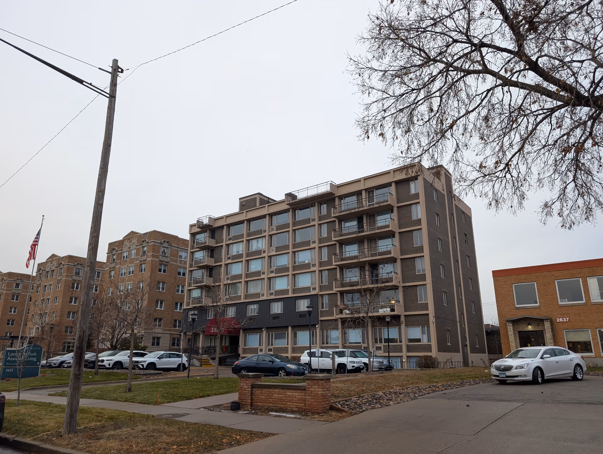 Six-story residential building with balconies, a small parking lot and an American flag under an overcast sky.
