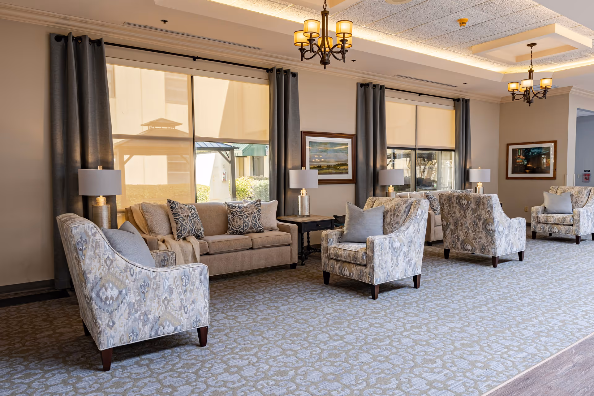 A spacious and well-lit common area in a senior living facility featuring multiple upholstered armchairs and sofas arranged around side tables with lamps. Large windows with dark curtains allow natural light to fill the room. The walls are decorated with framed landscape paintings, and elegant chandeliers hang from the ceiling.