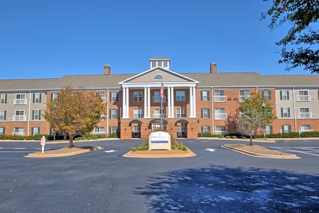 Front exterior view of a large three-story brick building with white columns and a central entrance. The building is surrounded by a parking lot with some trees and landscaping. The sky is clear and blue.