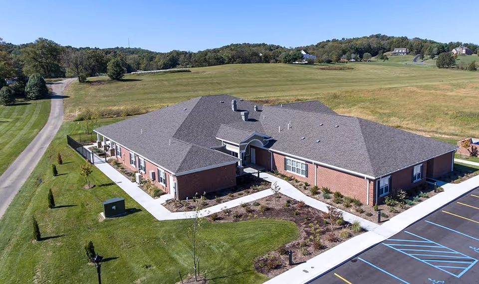 Aerial view of a single-story brick building with a gray roof, surrounded by green lawns and a parking lot with marked spaces. The building is situated in a rural area with open fields and scattered houses in the background.