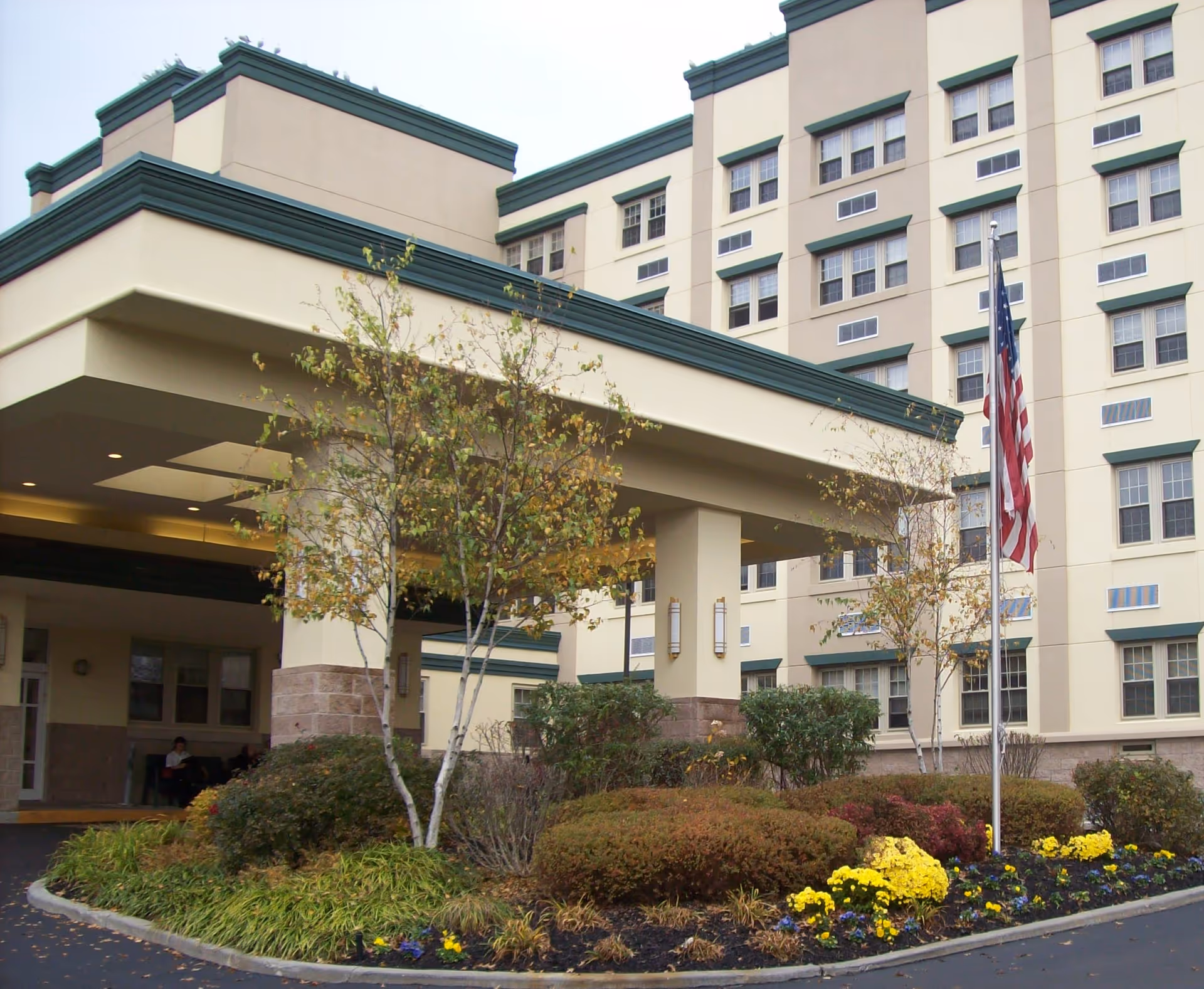 Front entrance and porte-cochere of a multi-story senior living building with landscaping and an American flag.
