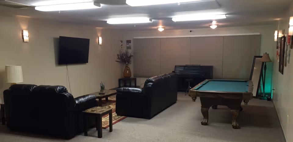 A common area with two black leather sofas facing a wall-mounted flat screen TV. There is a coffee table and side tables with lamps. To the right, there is a pool table and a piano against the back wall with closed blinds. The room is softly lit with ceiling lights and wall sconces.