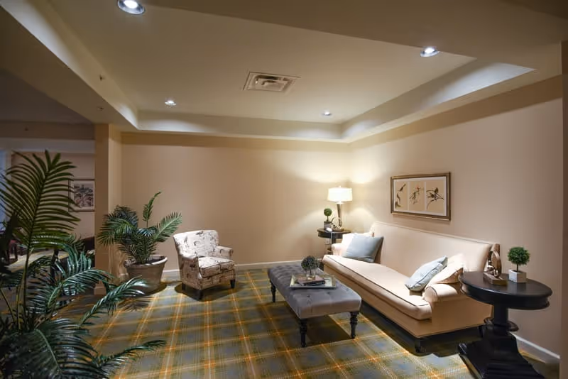 A cozy living room area with a beige sofa adorned with light blue and white pillows, a patterned armchair, a tufted ottoman serving as a coffee table with books and decorative items on it, two round side tables with small plants, a floor lamp, framed bird artwork on the wall, and several green potted plants. The room has a plaid carpet and recessed ceiling lights.