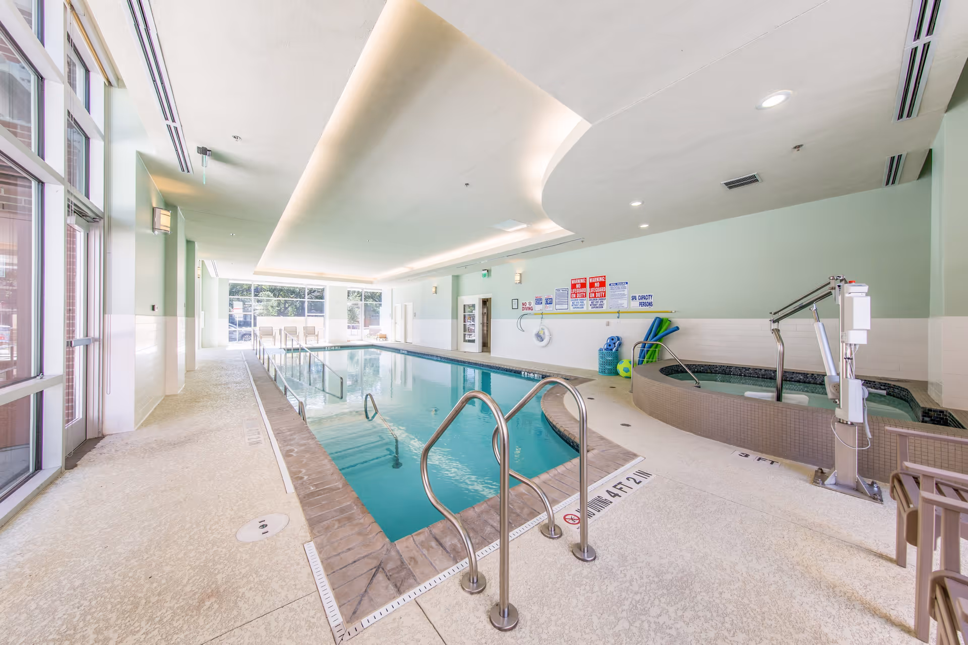 Indoor swimming pool area with a rectangular pool and a round hot tub. The pool has metal handrails and depth markers along the edge. The room has large windows on one side allowing natural light to enter. There are pool safety signs on the wall and pool equipment such as noodles and balls stored nearby. The ceiling has recessed lighting and a modern design.